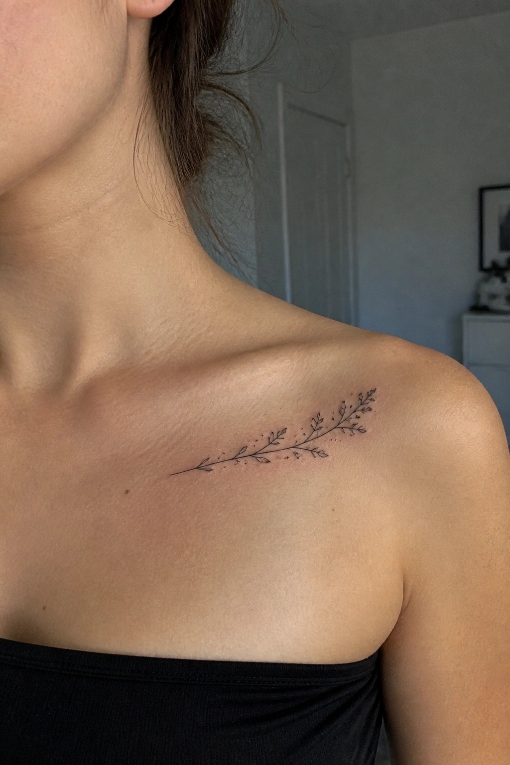 Bare shoulder with delicate black leafy branch tattoo above black strapless top