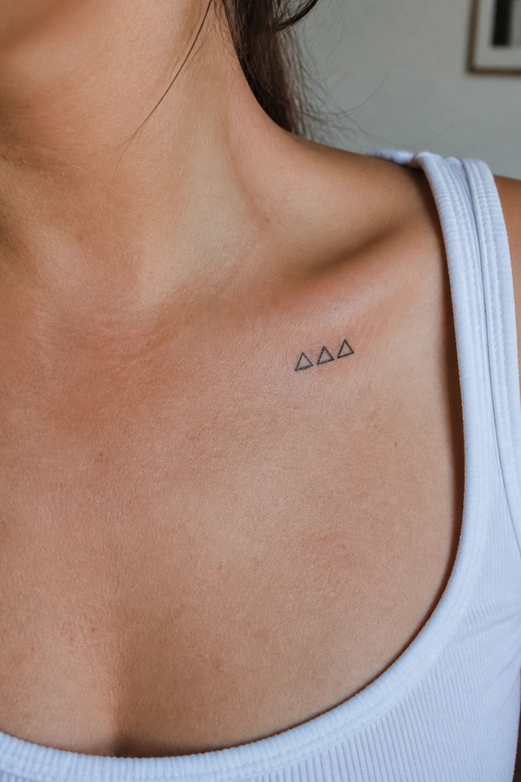 Three small black triangle tattoos on light skin above white tank top strap