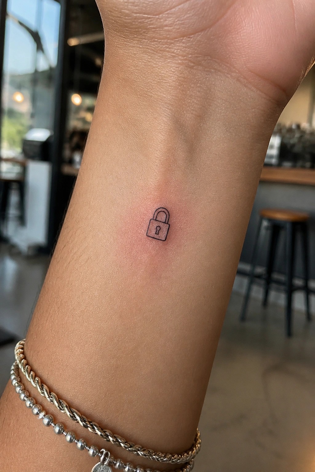 Small black padlock tattoo on wrist with silver beaded and braided bracelets