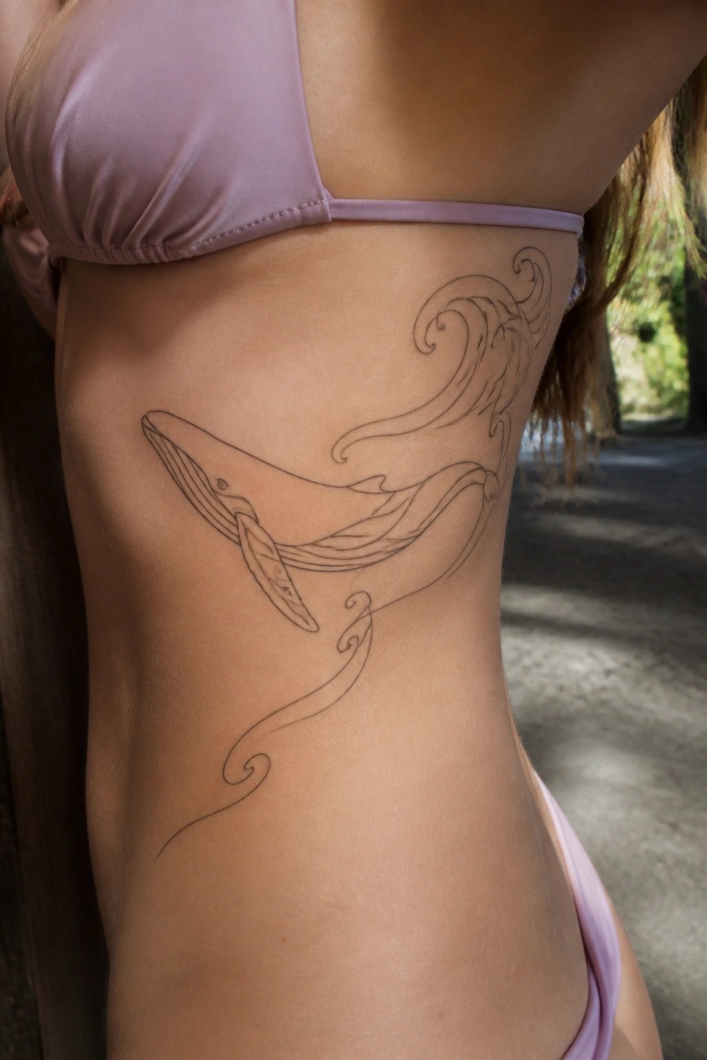 Woman's side with black line whale and waves tattoo in light purple bikini.