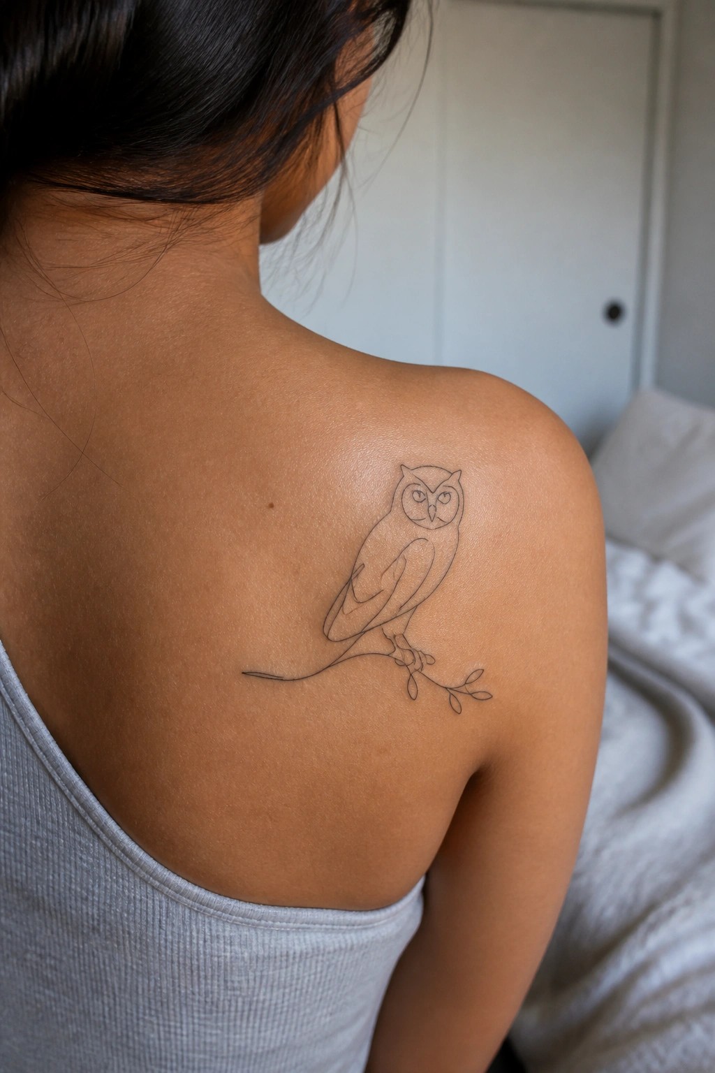 Woman's shoulder with minimalist owl line tattoo above gray tank top in bedroom