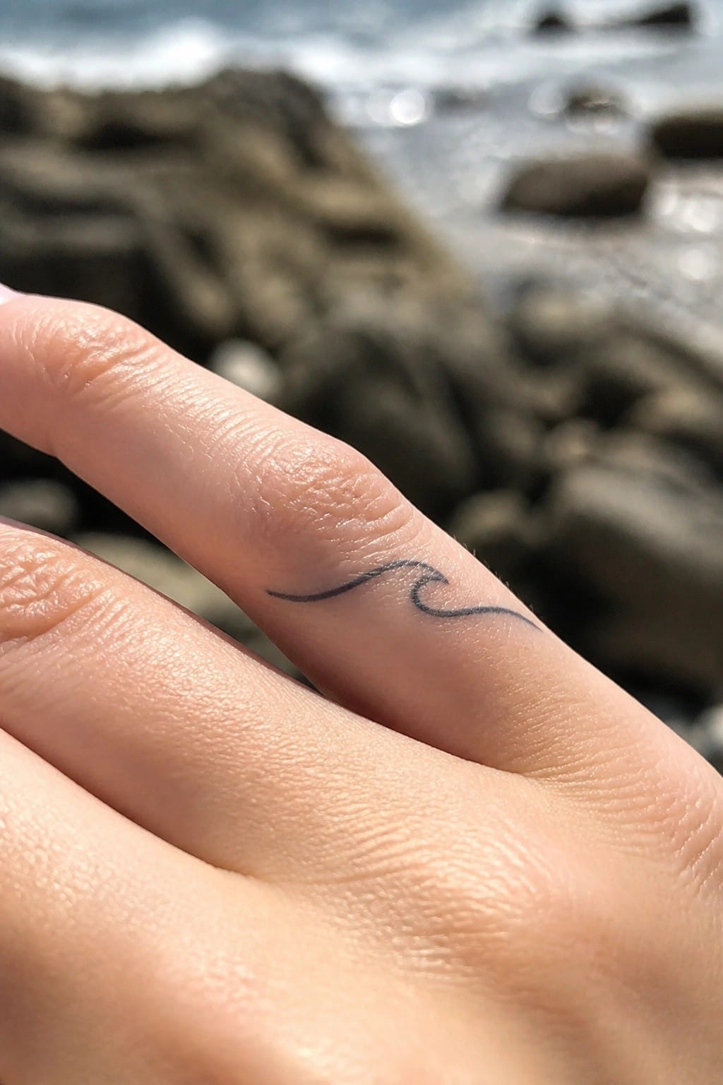 Close-up of light-skinned finger with small black wave tattoo and blurred ocean rocks behind