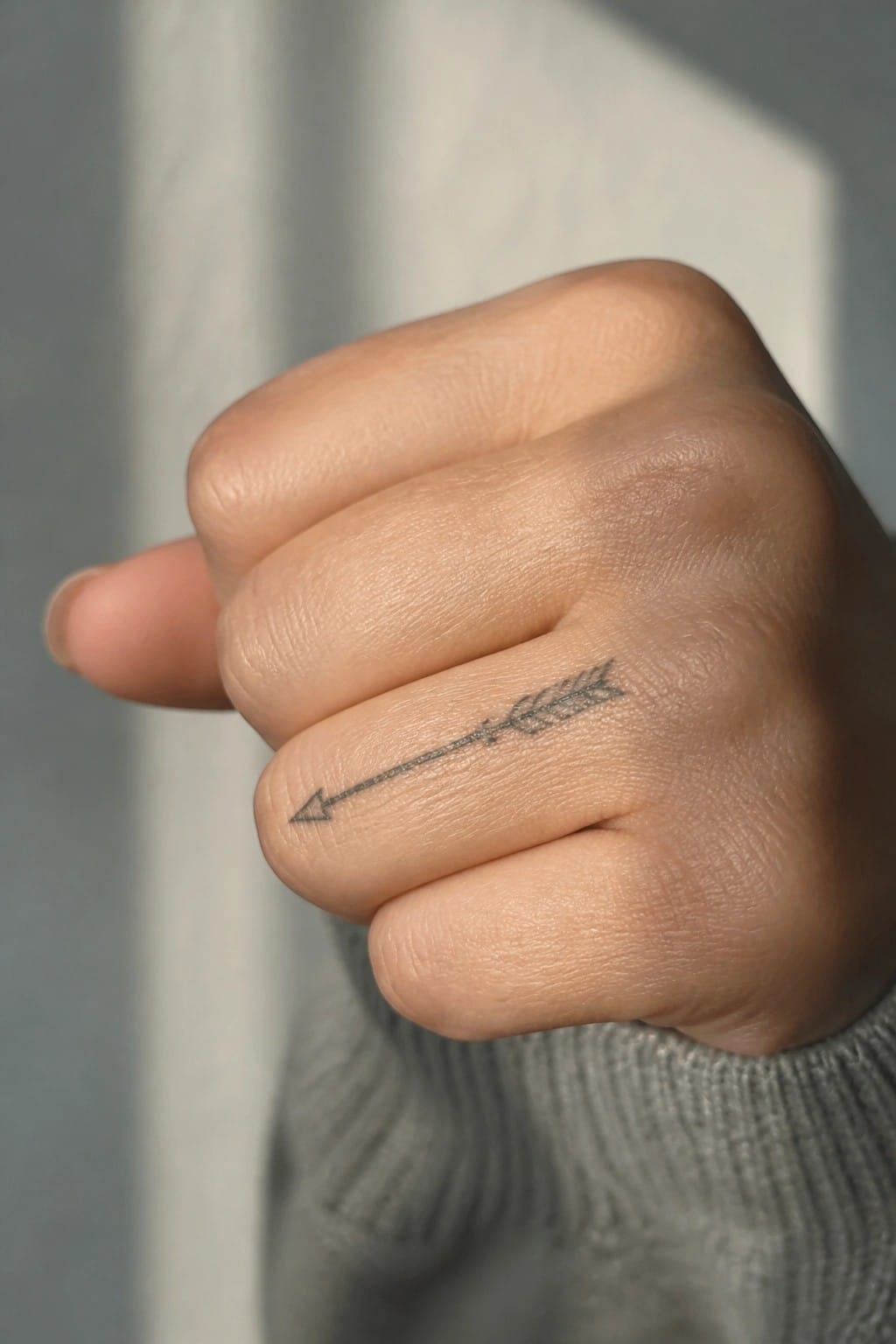 Small black arrow tattoo on the side of a finger, with the point facing left and simple fletching at the other end.