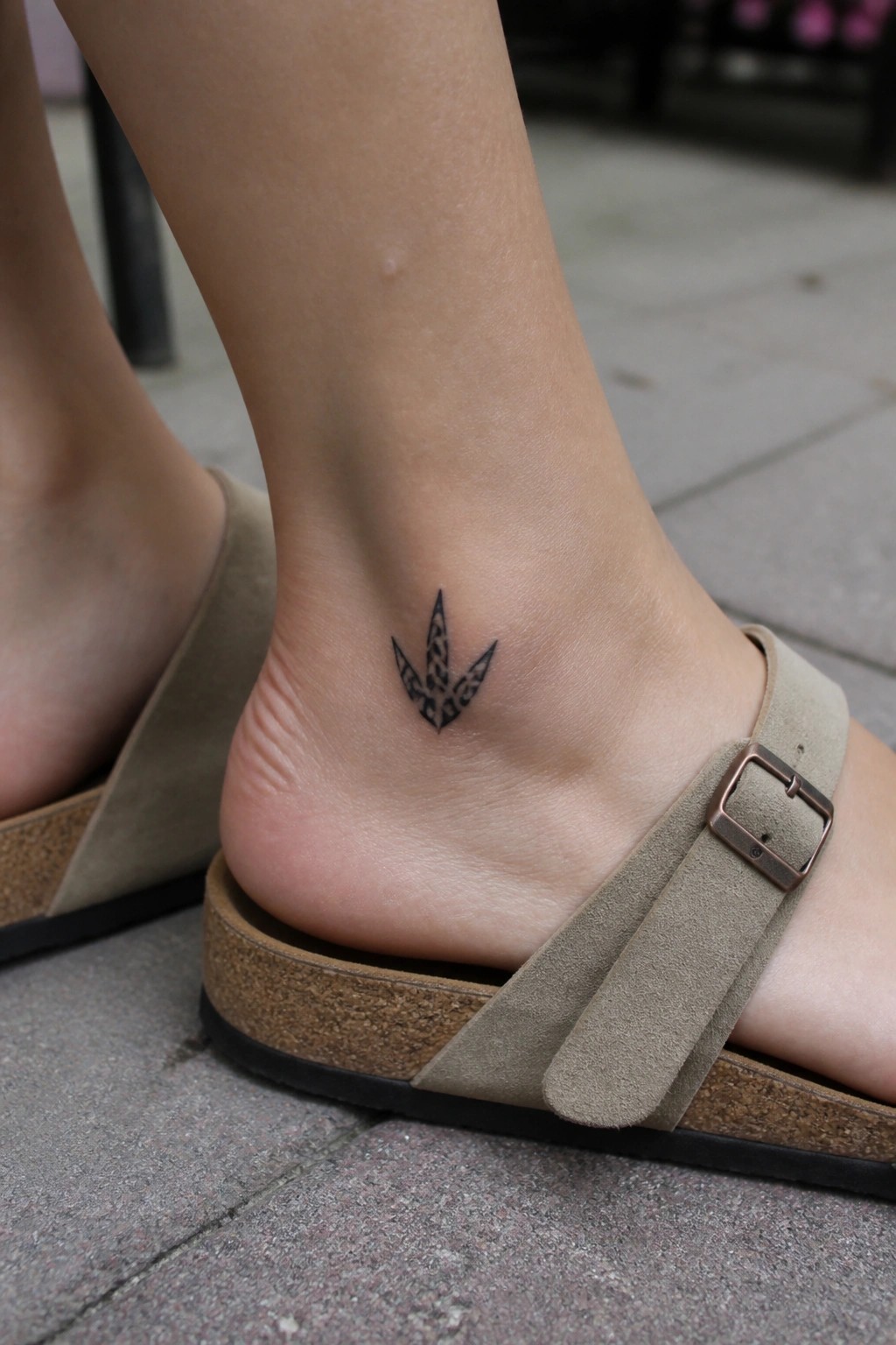 Black geometric tattoo on ankle above beige suede sandal with metal buckle on pavement.