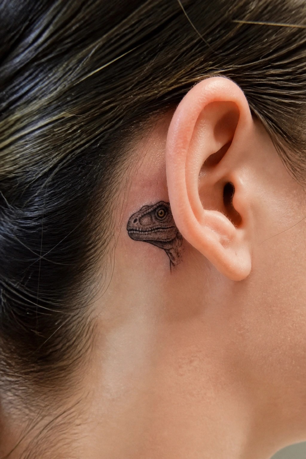 Small dinosaur tattoo with yellow eye behind ear amid dark hair