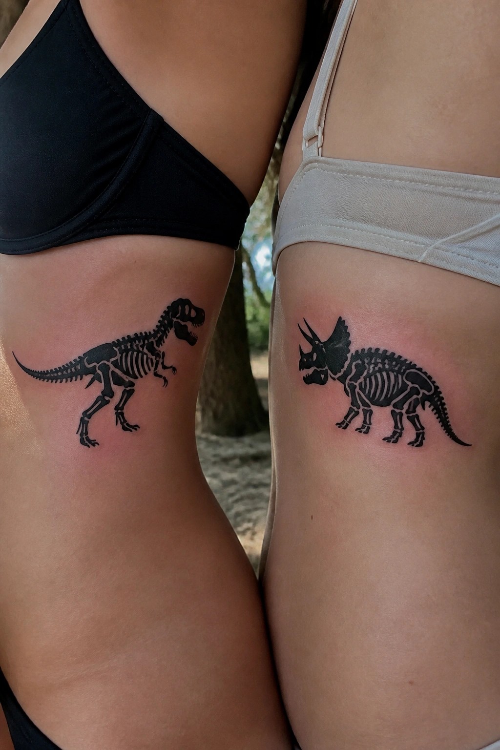 Two people display matching black dinosaur skeleton tattoos on their upper thighs, featuring a T-Rex on one side and a Triceratops on the other.