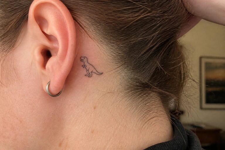 Minimalist black T-Rex tattoo behind ear with diamond stud earring in brown hair