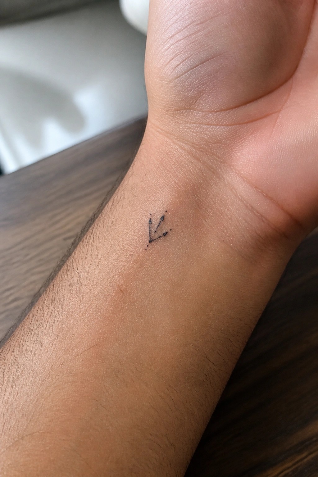 Small black arrow tattoo on light-skinned wrist above wooden surface.