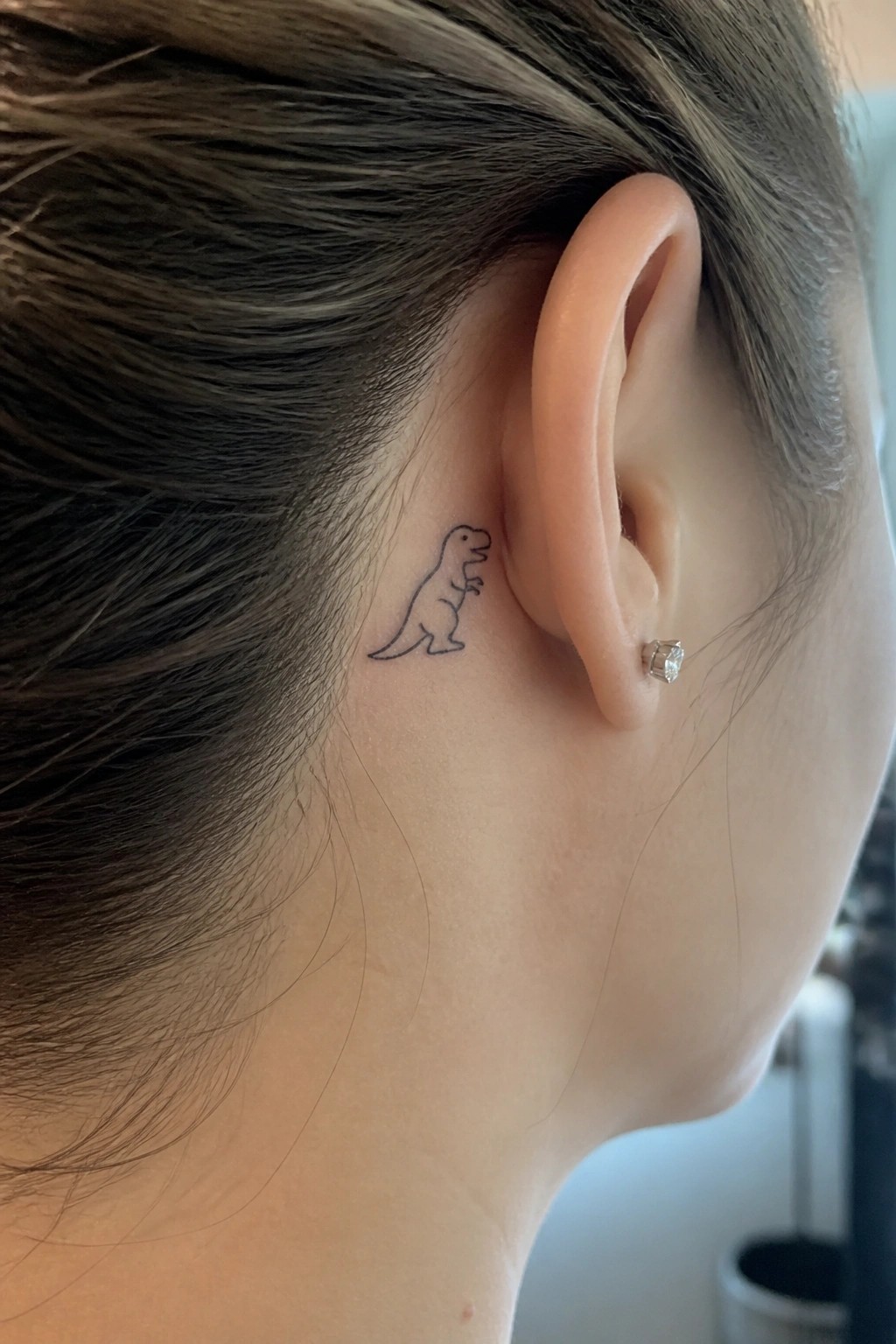 Minimalist black T-Rex tattoo behind ear with diamond stud earring in brown hair