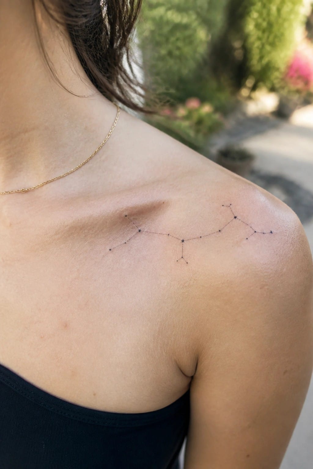 Shoulder with constellation tattoo, gold necklace, black top, blurred garden background