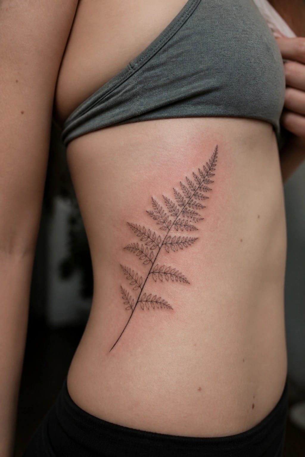 Fine line fern tattoo on the side of the torso showing the full stem and leaves in black ink
