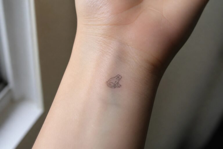 Small minimalist frog tattoo on inner wrist of light-skinned hand