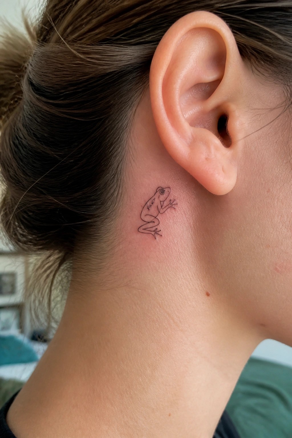 Small black frog tattoo behind the ear of a person with brown hair in a bun.