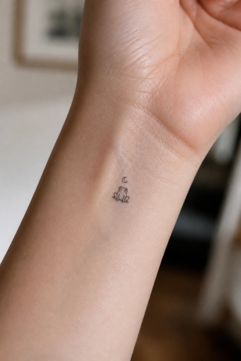 Minimalist black line tattoo of frog under crescent moon on inner wrist.