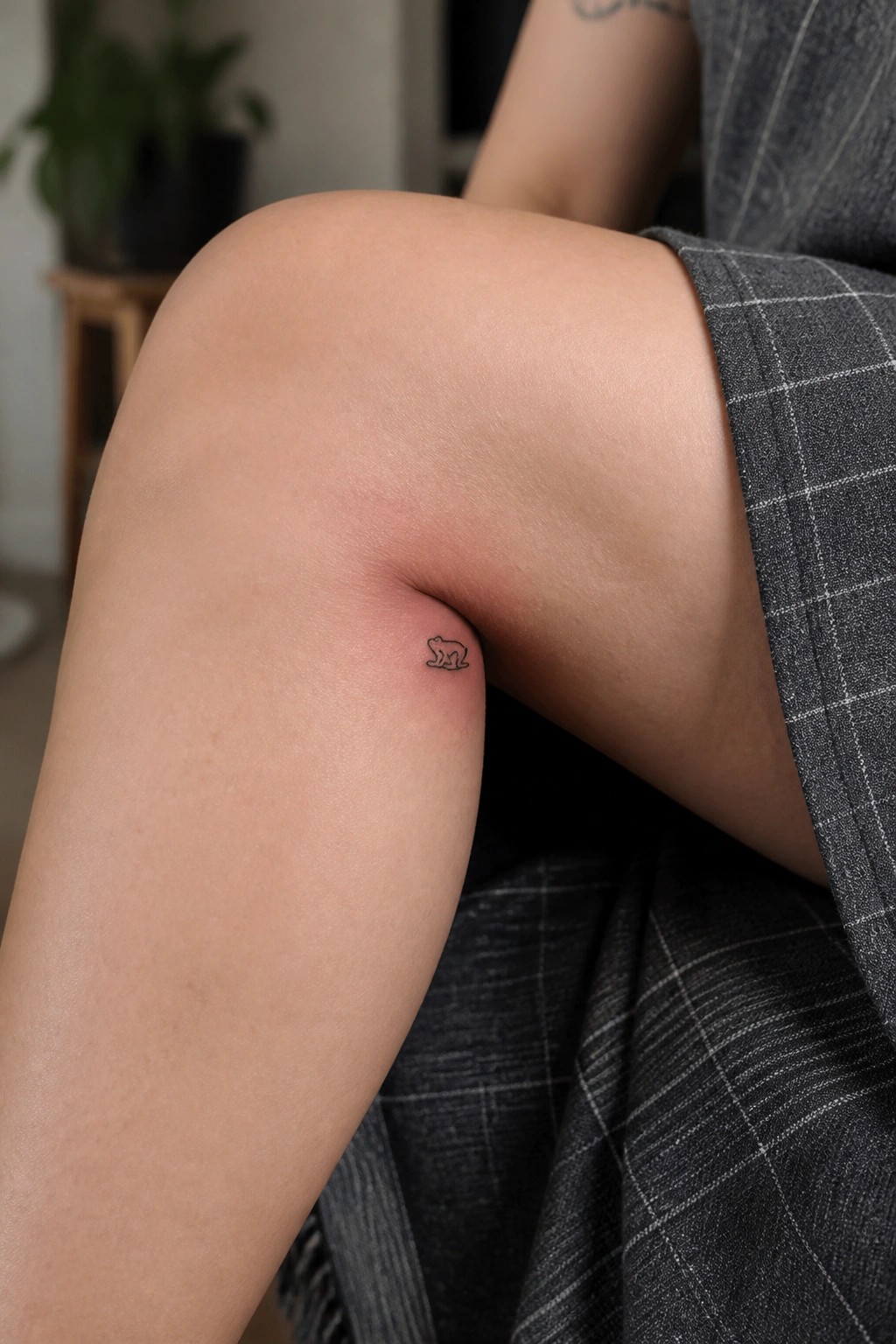 Small frog tattoo on reddened skin beside gray plaid fabric on bent knee.