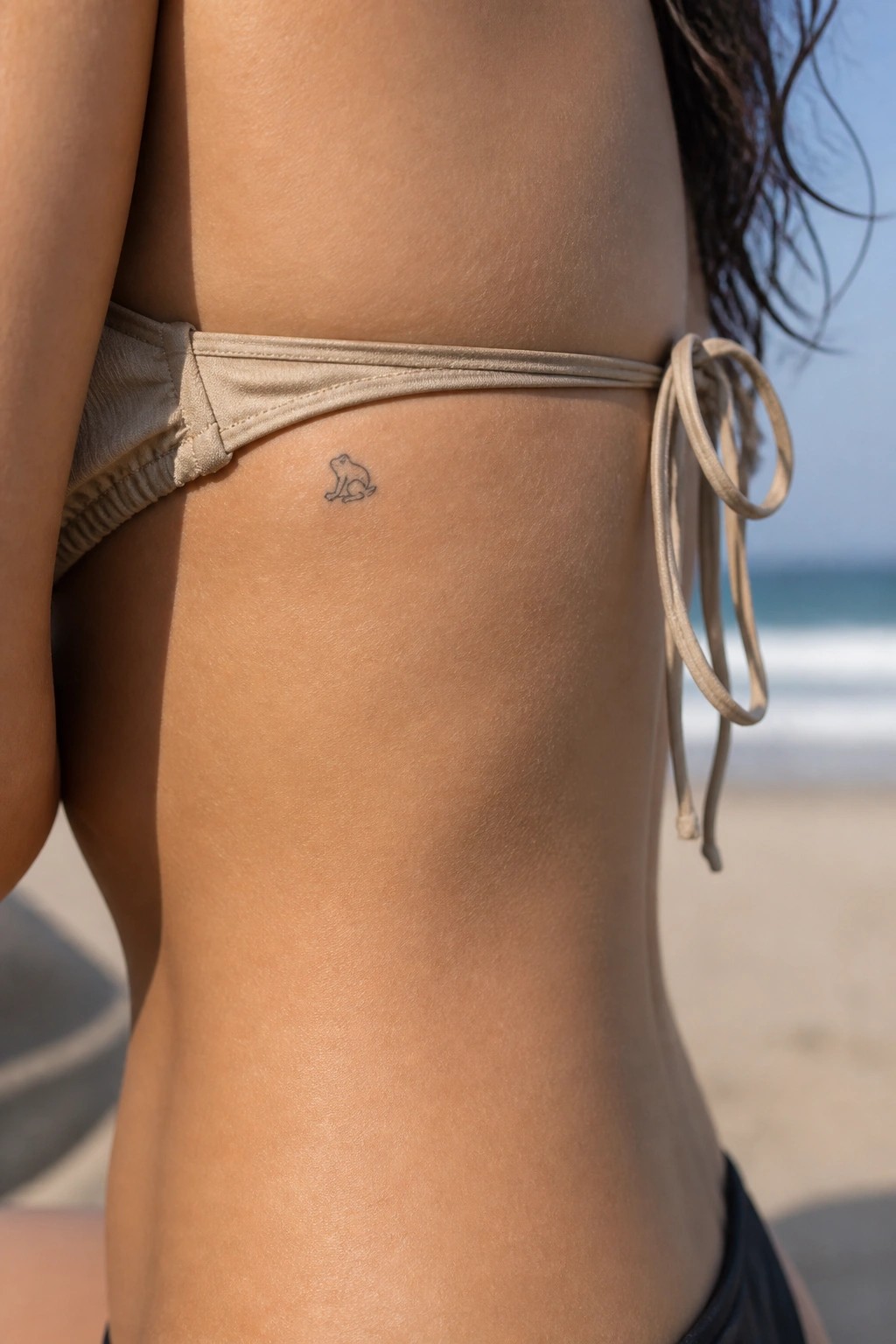Tiny frog tattoo on hip above tied beige bikini at sunny beach