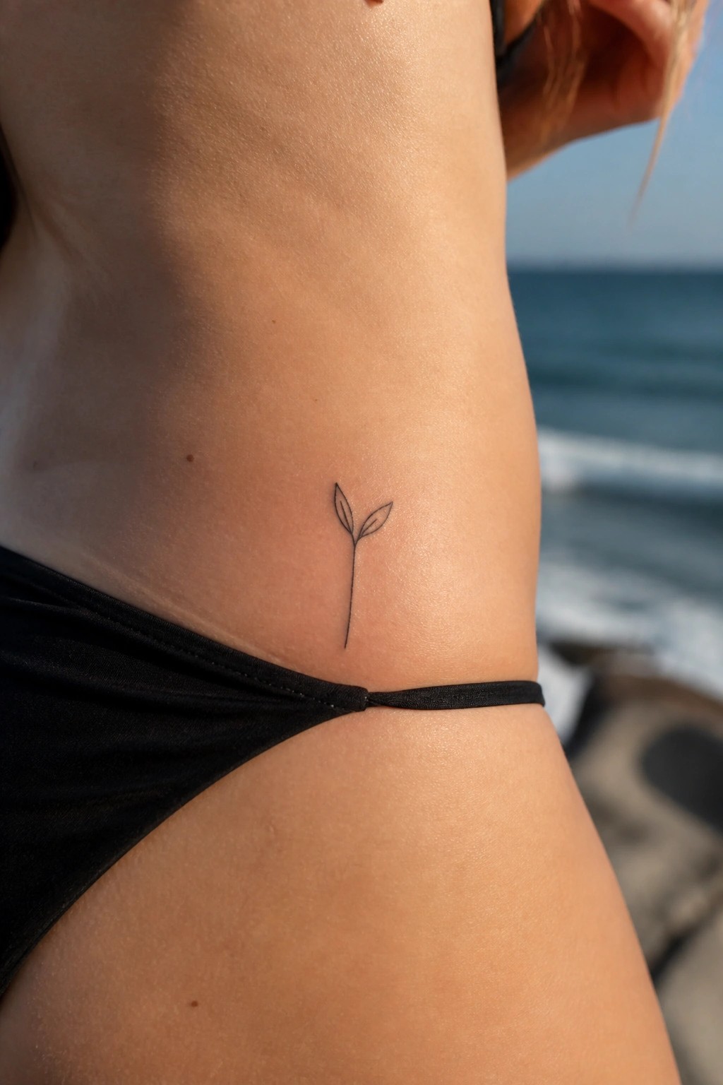 Small plant sprout tattoo on hip above black bikini with ocean background