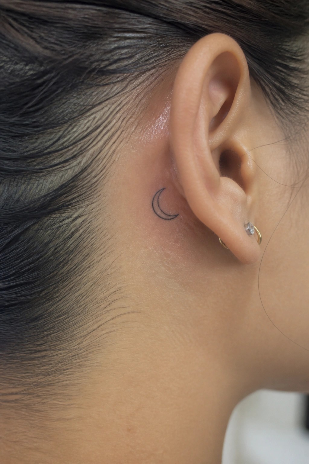 Side view of ear with crescent moon tattoo and small gold earrings.