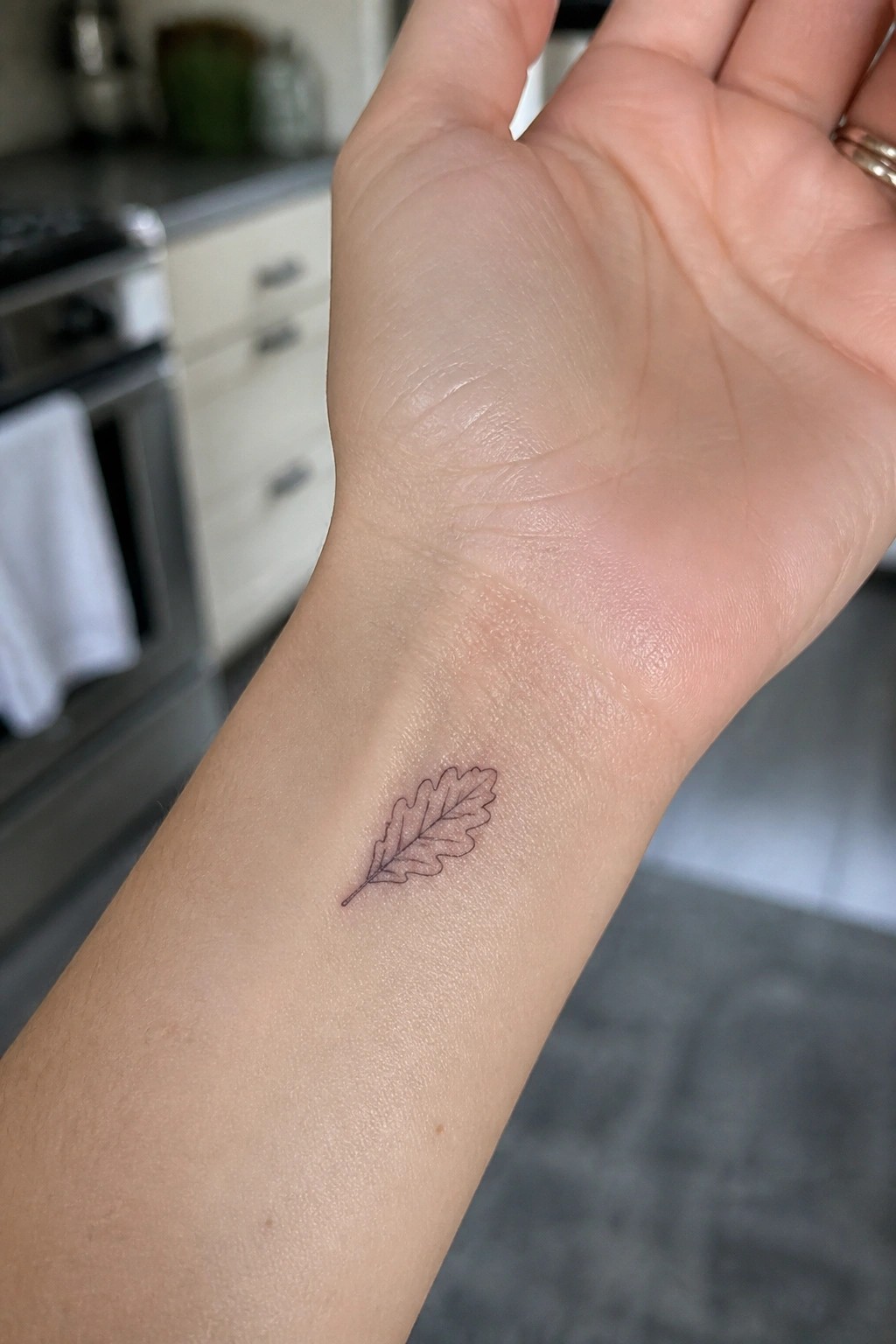 Wrist with fine-line oak leaf tattoo in blurred kitchen background.