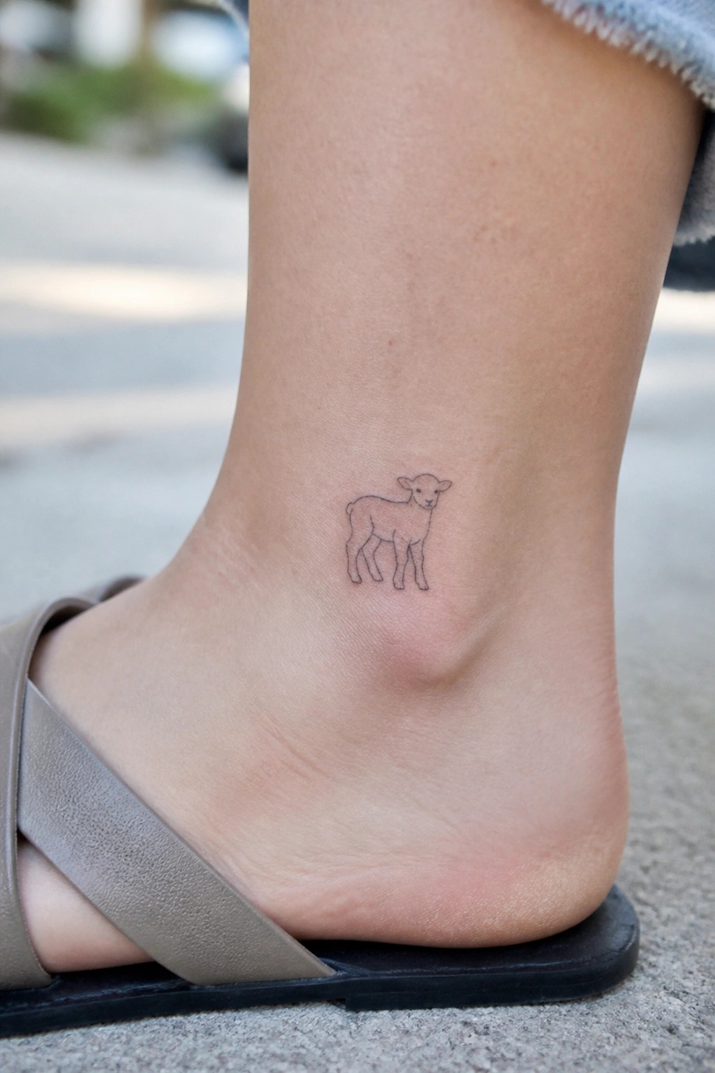 Small lamb outline tattoo on ankle above beige sandal strap on pavement