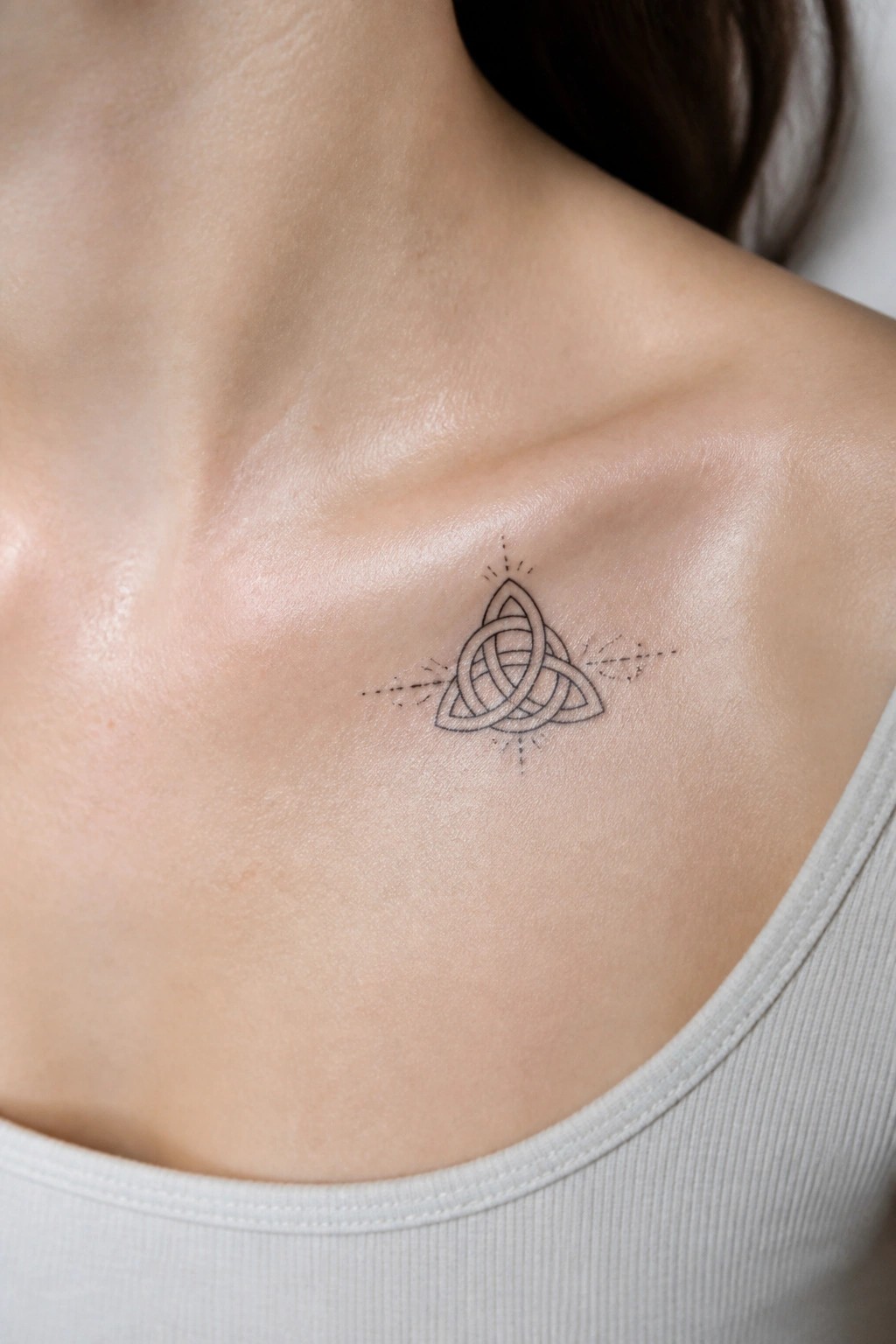 Minimalist Celtic knot tattoo with dotted rays on collarbone over light tank top.