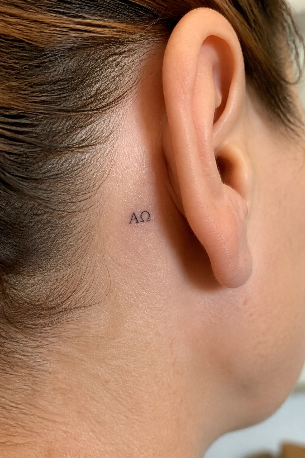 Small black AΩ tattoo behind a person's ear on light skin with brown hair