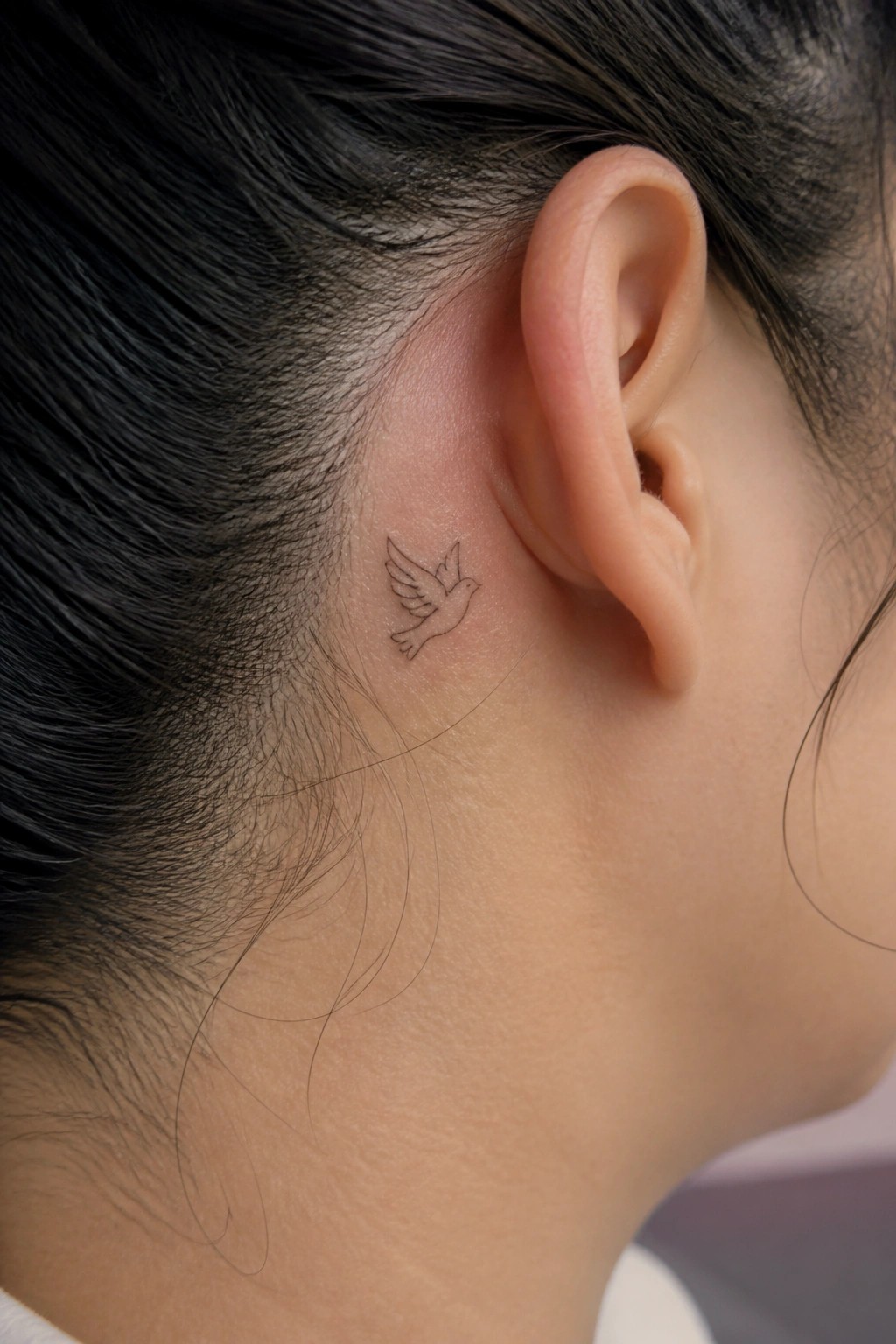 Small bird tattoo behind ear on dark-haired person with pulled-back hair.
