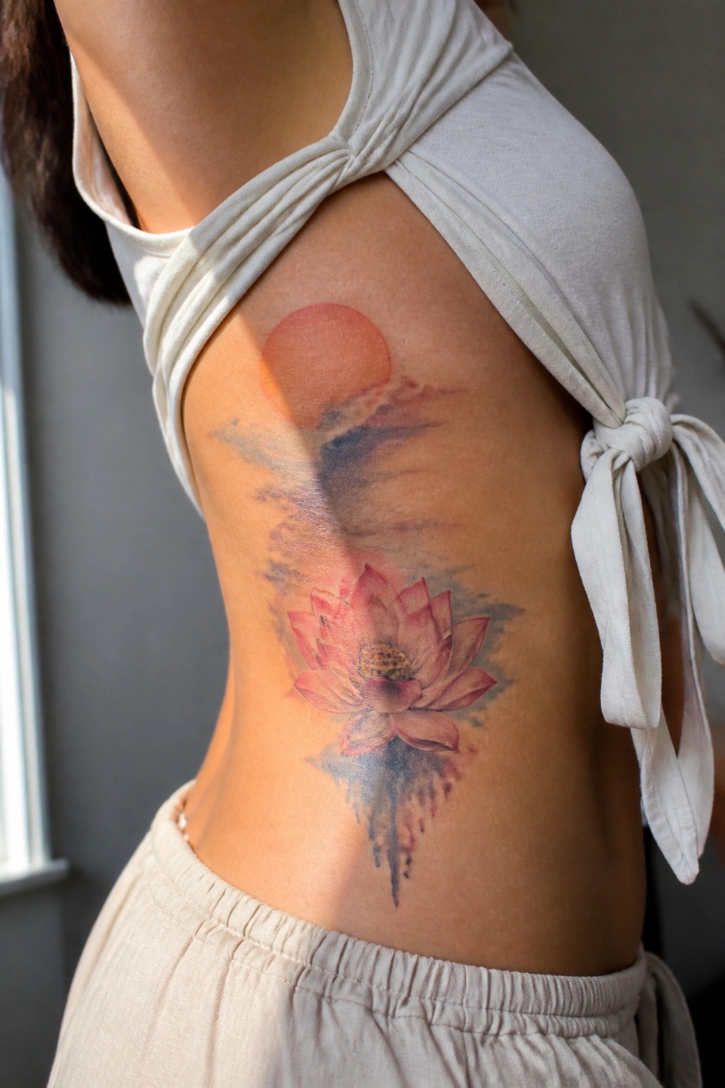 Watercolor lotus tattoo with red sun and clouds on woman's side in white tied top.