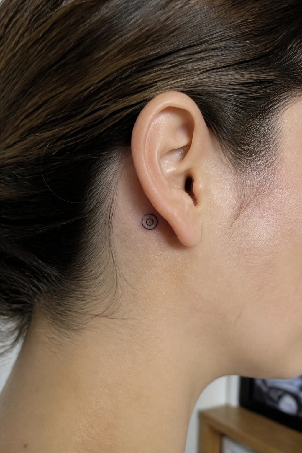 Small black spiral tattoo placed just behind the ear on the side of the head.