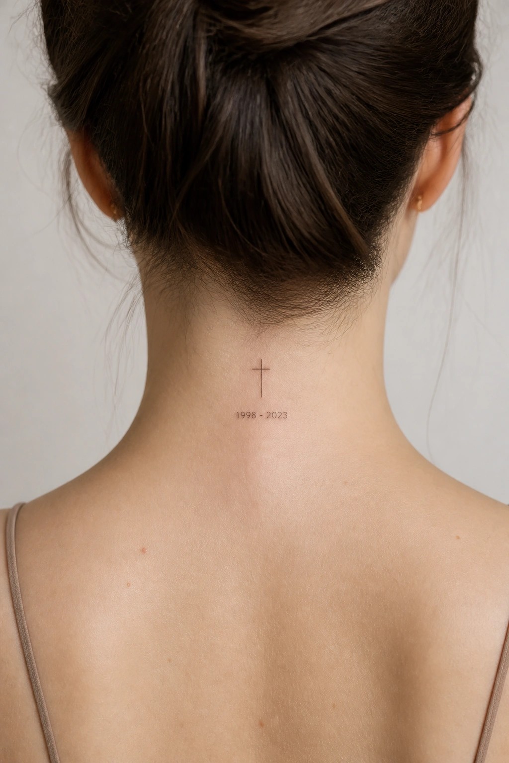 Small cross tattoo with dates 1998-2023 placed on the nape of the neck, shown from behind with hair in a bun.