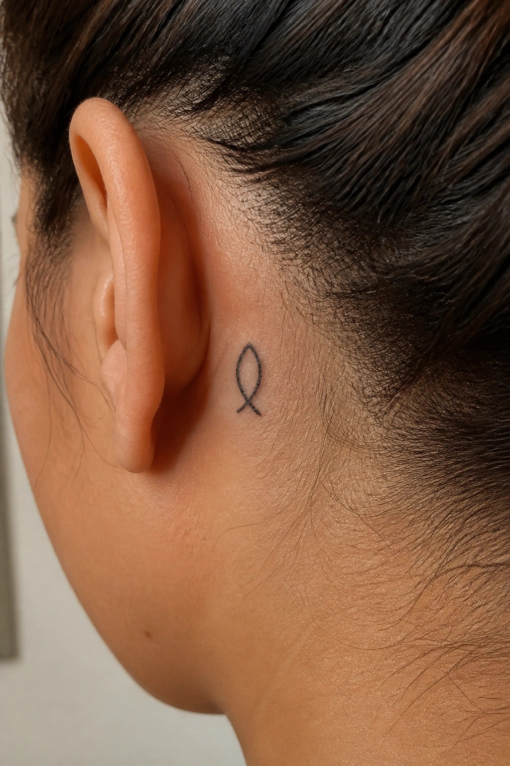 Small black outline of a Christian fish symbol tattooed behind the ear on a person's head.