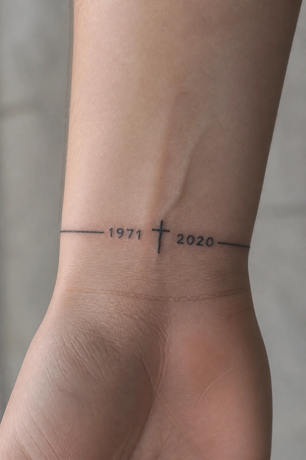 Minimalist wrist tattoo with a thin horizontal line, the years 1971 and 2020, and a small cross centered between them.