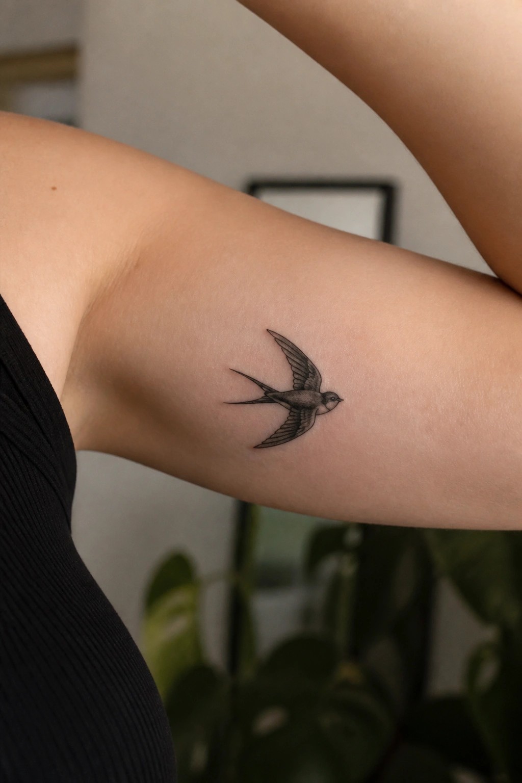 Black swallow tattoo on upper arm of person in black sleeveless top.