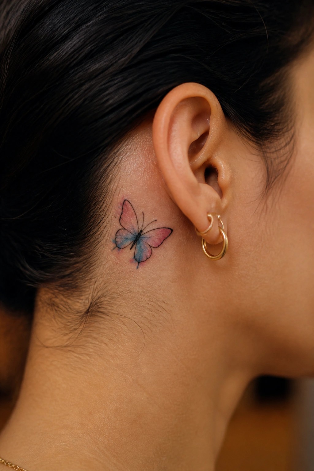 Watercolor pink and blue butterfly tattoo behind ear with gold hoop earrings.