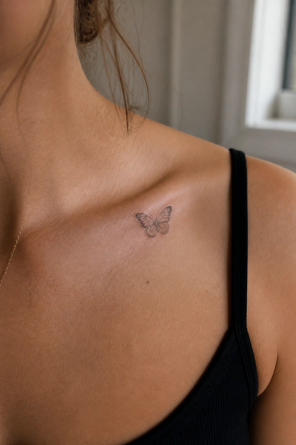 Small butterfly tattoo on collarbone above black tank top strap with gold chain
