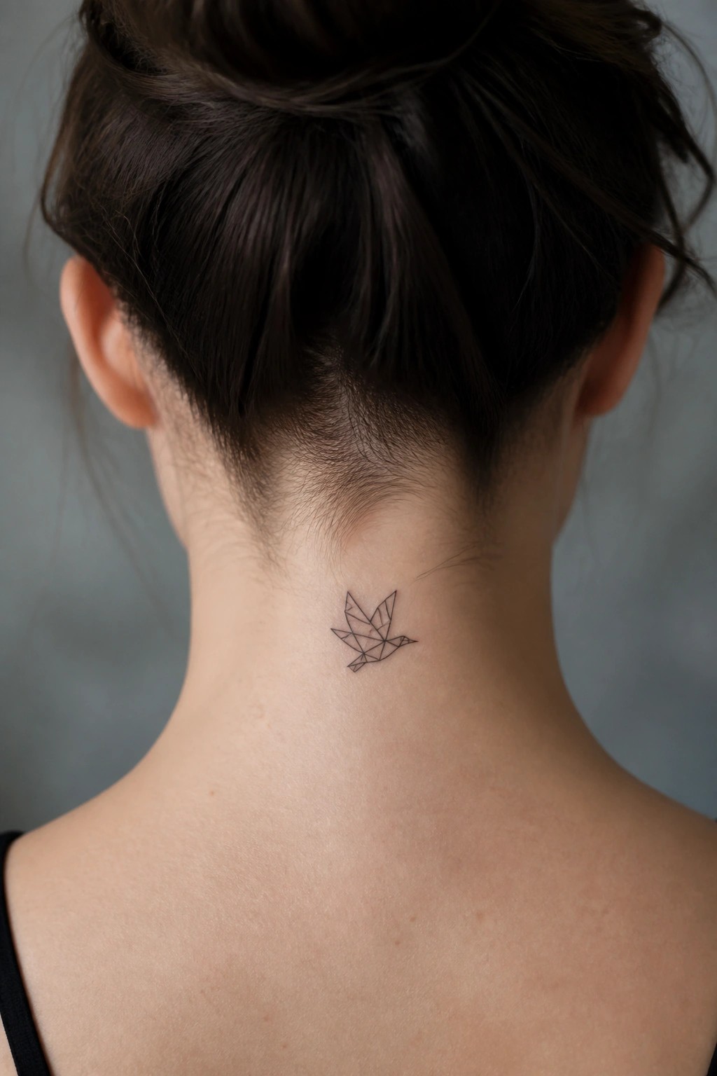 Woman's neck with small geometric bird tattoo below dark hair bun.