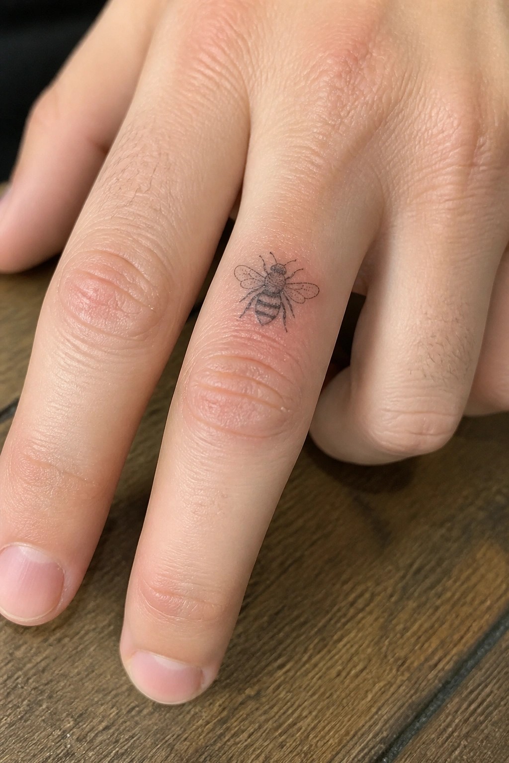 Small black bee tattoo on a finger resting on wooden surface
