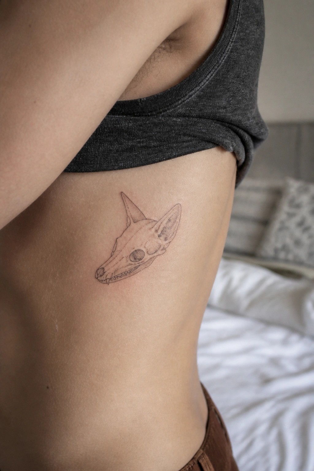 Line-art fox skull tattoo on side under lifted gray tank top in bedroom.