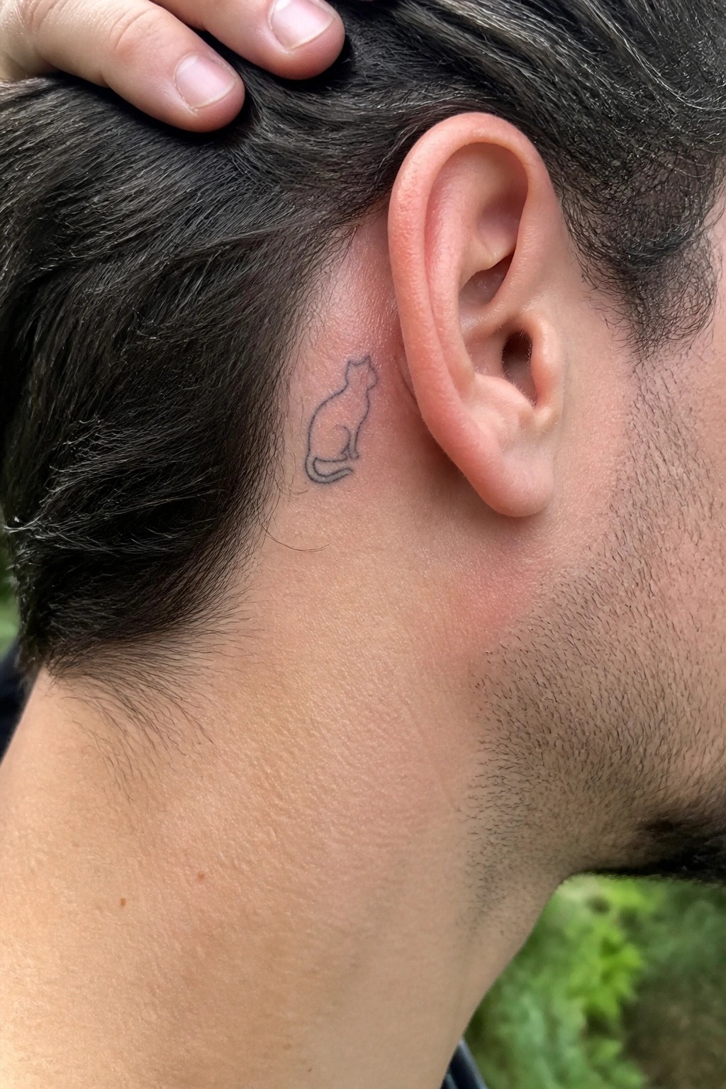 Small cat tattoo behind ear, hand resting on dark hair.