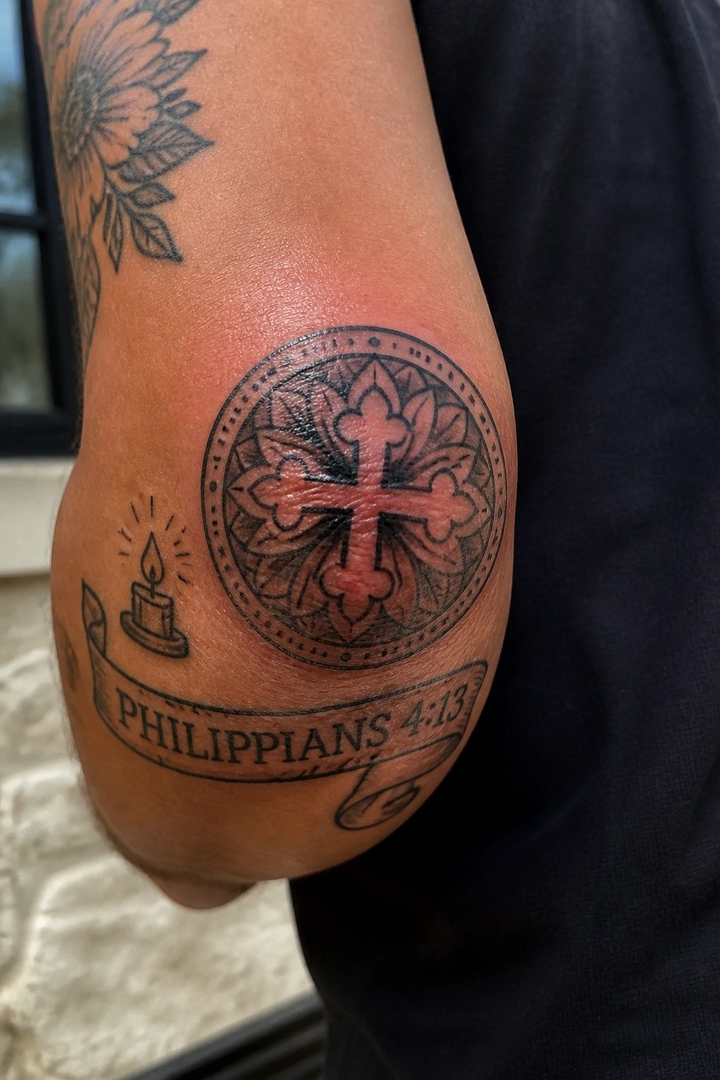 Elbow tattoo of a cross inside a circular frame with a Philippians 4:13 banner and small candle element below
