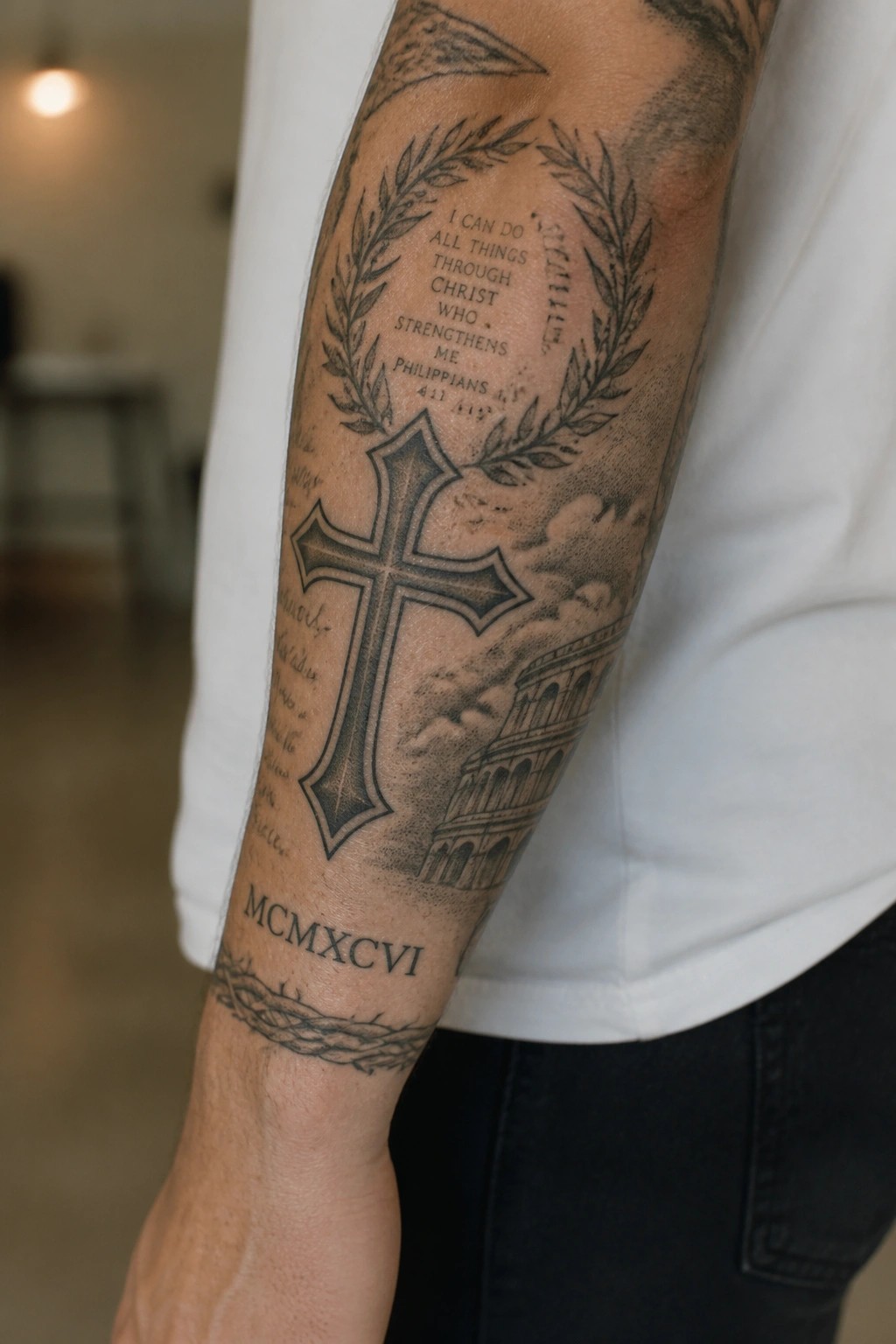 Black and grey forearm tattoo with a central cross, laurel wreaths, Philippians 4:13 verse, and Colosseum illustration.