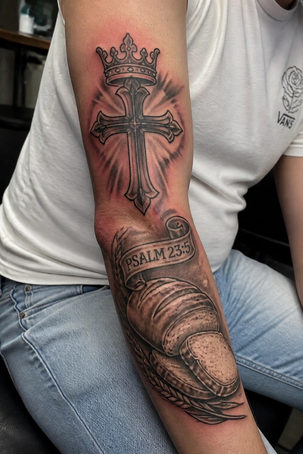 Arm tattoo of crowned cross above bread loaf with Psalm 23:5 banner on white shirt