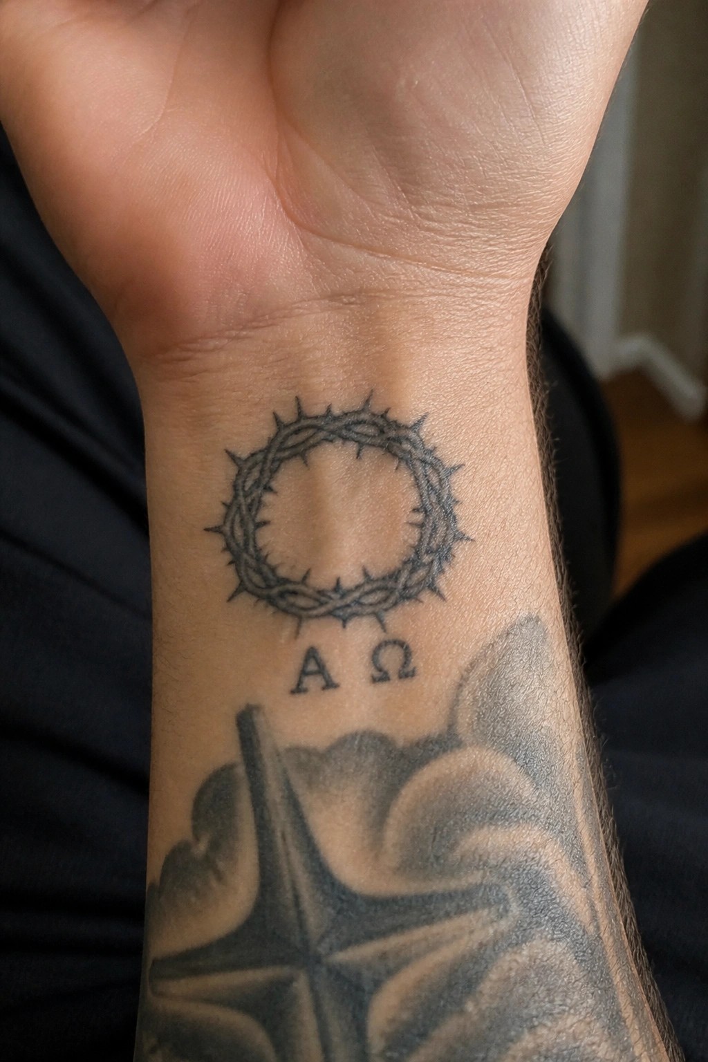 Crown of thorns tattoo with A Ω letters on wrist above cross and star.