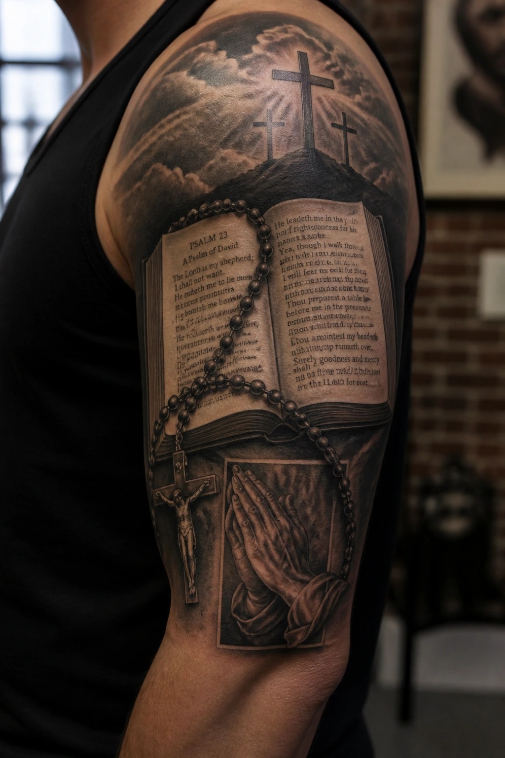 Black and gray realistic tattoo of an open Bible showing Psalm 23, three crosses on a hill, rosary beads, praying hands, and a crucifix covering the upper arm and shoulder.