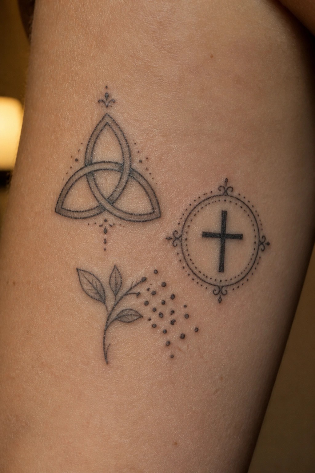 Fine line tattoo of a triquetra, a circular cross emblem, and a small leafy plant on the upper arm