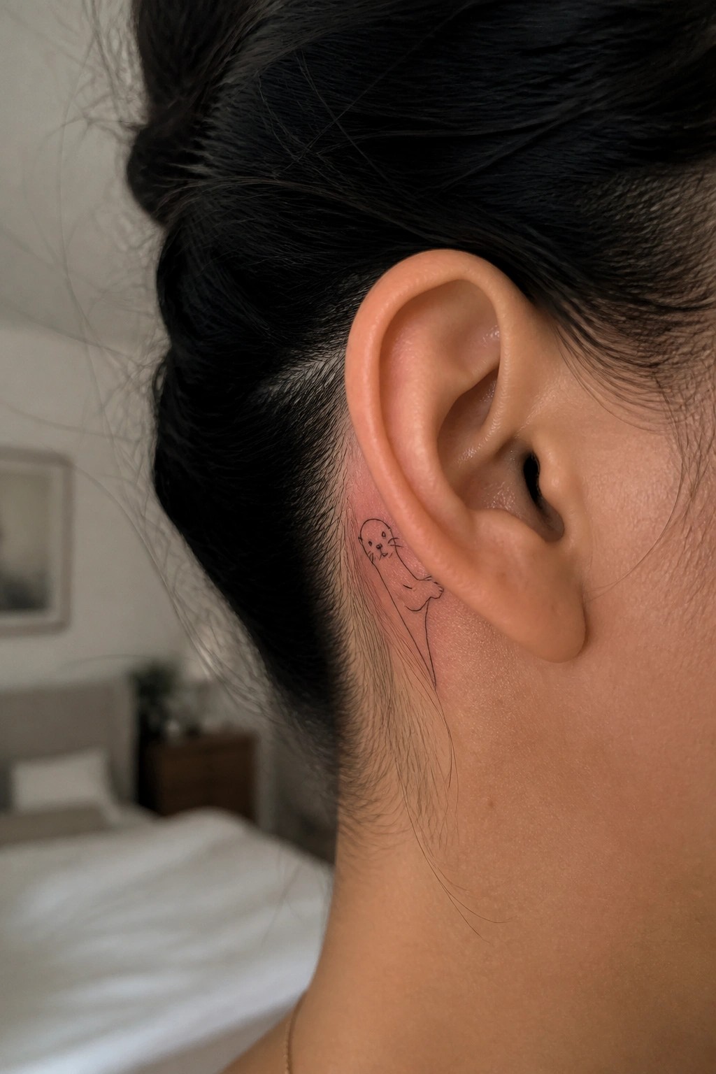 Minimal black line tattoo of a small otter placed directly behind the ear on the side of the head.