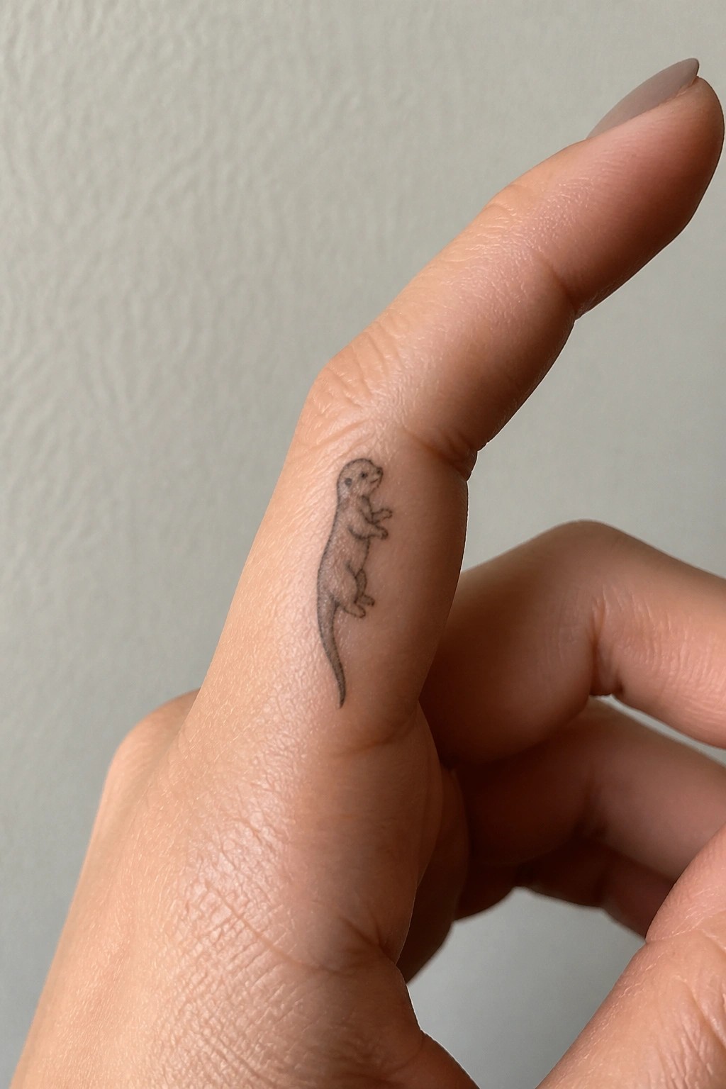 Minimal black line otter tattoo standing on the side of a finger with tail extending downward.