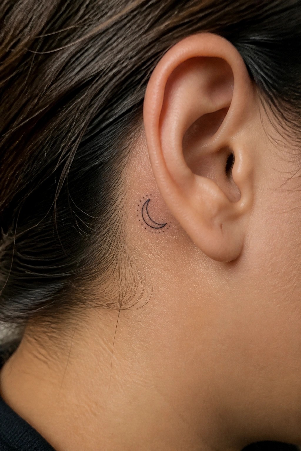 Crescent moon tattoo with dots behind a person's ear on light skin.