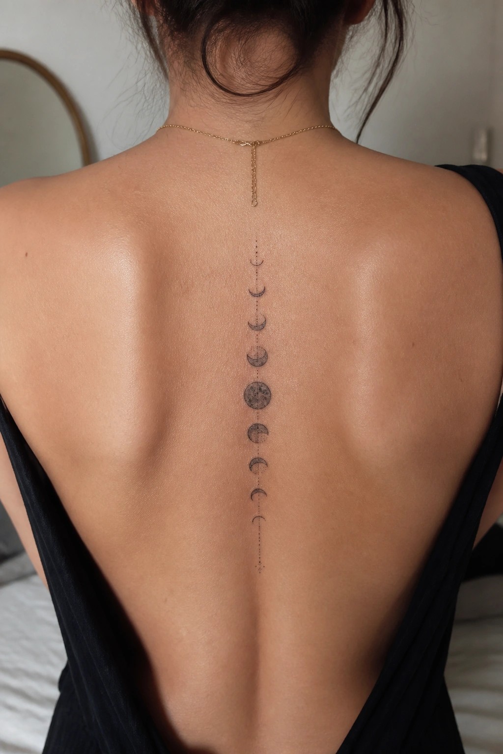 Woman's back with moon phases tattoo, gold necklace, black open-back top