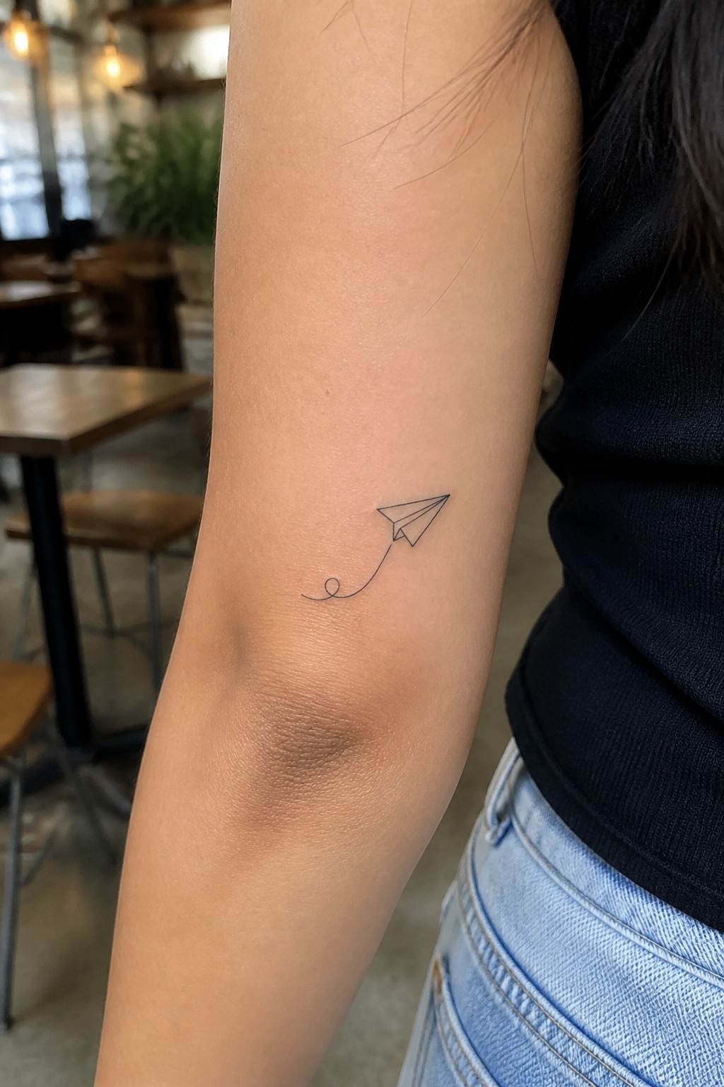 Fine line paper airplane tattoo on the upper arm with a trailing loop detail